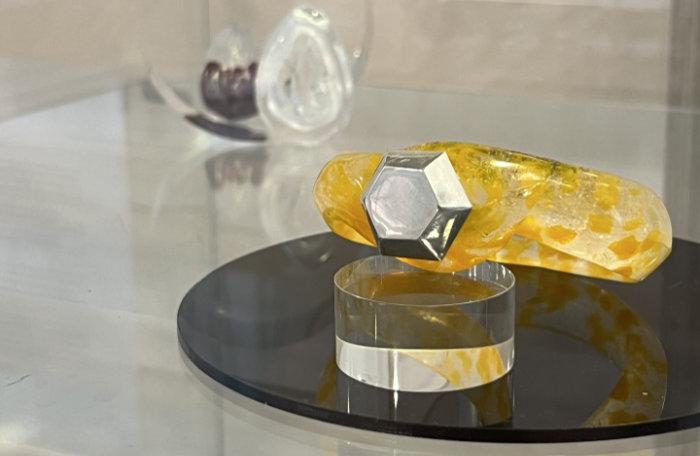 A chunky solid band bangle made with faceted yellow glass with a hexagonal mirrored glass feature detail displayed on a round perspex plinth. Small glass objects blurred in the background.