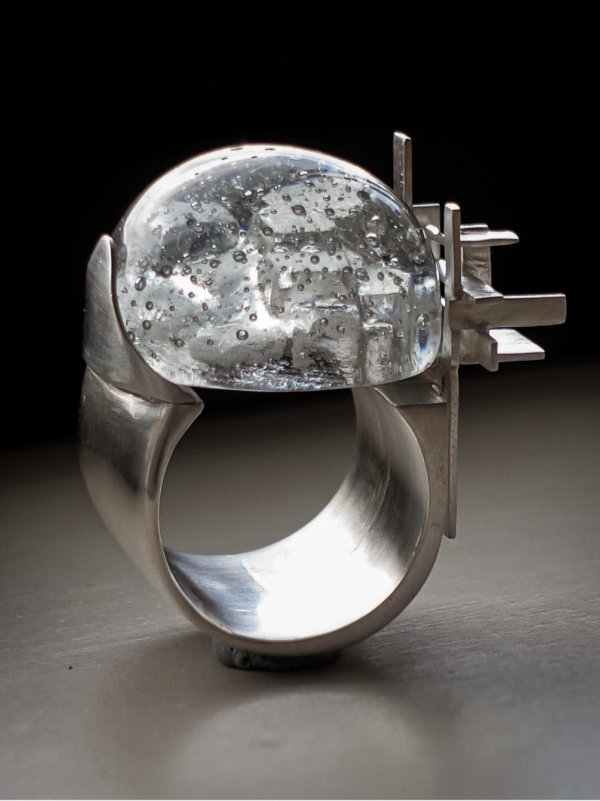 A wide-band silver ring shown side-on with a tiny cityscape on the top and jutting angular silver pieces going vertically and horizontally on one side and a greyish glass cabochon mounted over like a dome on the cityscape.