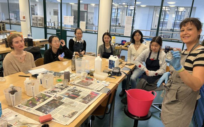 Mirroring demonstration with Joanna holding up a piece of mirrored glass on the right and students behind sitting round a large table to view it. A bucket sits on a stool and various bottles of solutions sit on newspaper on the table.