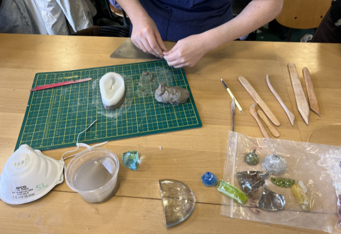 Overhead shot of table with tools and materials showing student's hands working on making an investment mould ready for jewellery making.