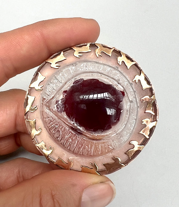 Round 'Rabbit Eye' brooch featuring a dark red glass blob in the centre and clear textured glass surrounding it and a ring of copper rabbit silhouettes running around the outside edge as part of the mount.