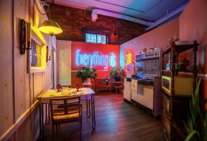 An image of a dimly lit kitchen scene with the word Everything in blue neon on the back wall against a red background. Title of piece is The Maiden Crone, Past, by Alleson Buchanan.