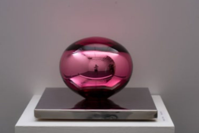 A horizontal egg-shaped piece of glass in ruby red standing on a narrow grey rectangular stand. The piece is by Tom Bennett and called Desire(I, Pink).